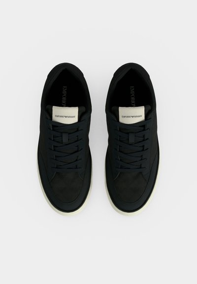 Paire de baskets en cuir noir Emporio Armani avec semelles blanches et étiquettes de languette à la marque, vues de dessus sur fond blanc.