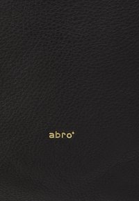 Surface en cuir noir texturé avec un petit logo de marque "abro" en or centré dans la moitié inférieure.