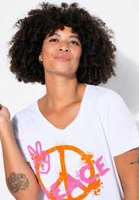 T-shirt blanc en V avec un symbole de paix orange vif et le texte "PEACE" en rose, présentant des accents de éclaboussures de peinture. Tissu en coton doux.
