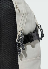 Le sac à dos présente un panneau en maille noire et un corps en toile beige clair, avec des cordons à motifs gris et noirs et un fermoir en plastique.