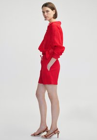 Mono de tela arrugada roja con un top corto abotonado, mangas largas, cintura con cordón y bolsillos laterales, combinado con tacones marrones de tiras.