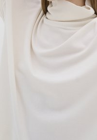 Biała sweter o miękkim, luźnym fasonie z wysokim kołnierzem oraz gładką fakturą. Wyraźne fałdy podkreślają jego swobodny krój oraz nowoczesny wygląd.