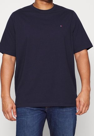 Hombre vistiendo una camiseta de manga corta azul marino con un pequeño logo rojo, blanco y azul en el pecho, combinada con jeans azules.