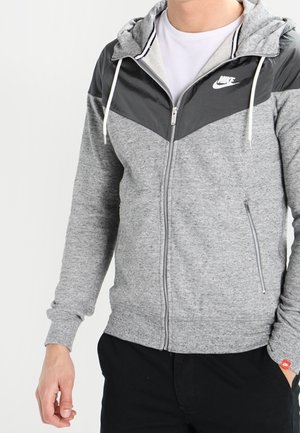 Mann trägt einen grauen Nike Zip-up Hoodie mit schwarzem Chevron-Detail über der Brust und weißen Kordeln, kombiniert mit schwarzen Hosen.