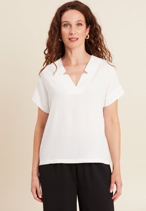 Donna con capelli ricci castani che indossa orecchini a cerchio dorati, una camicetta bianca a maniche corte con scollo a V e pantaloni neri, in piedi davanti a uno sfondo neutro.