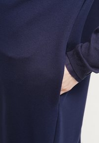 Mörkblå, mjuk klänning i tyg med en sidficka, långa ärmar med ribbad manschett och en slät, elegant yta.