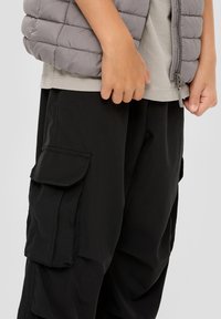 Grau gesteppte Steppweste, getragen über einem hellgrauen T-Shirt. Schwarze Cargo-Hose mit mehreren Taschen, die eine glatte Textur aufweist.