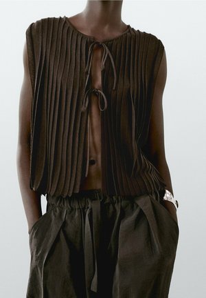 Personne portant un gilet plissé sans manches marron foncé noué avec deux cordons à l'avant, avec un pantalon ample assorti et un bracelet en argent au poignet.
