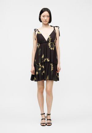 Kobieta o krótkich czarnych włosach, ubrana w czarną sukienkę bez rękawów z żółtymi kwiatowymi wzorami i czarne sandały na paski, stojąca na tle jednolitego tła.