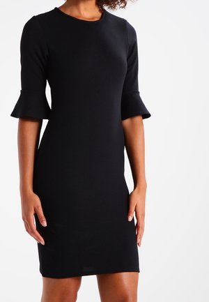 Femme portant une robe noire cintrée longueur genou avec des manches à volants jusqu'aux coudes sur un fond blanc uni.