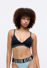 Bralette triangle noire avec des straps fins, présentant une bande logo grise. Culotte bleu clair avec une ceinture logo noire. Texture de tissu lisse.