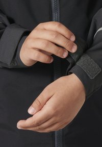 Czarna kurtka z zamkiem, z regulowanymi mankietami na rzep. Zbliżenie na ręce dostosowujące rękaw, ukazujące gładką teksturę materiału.