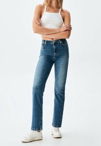 Mujer de pie con los brazos cruzados, vistiendo una camiseta corta blanca, jeans de tiro alto azul y zapatillas blancas sobre un fondo blanco liso.