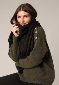 Écharpe noire avec un motif en chevrons, ornée de fils métalliques subtils, drapée autour d'un pull vert avec des accents de boutons sur l'épaule.