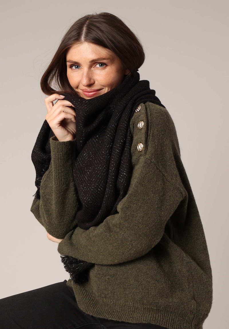 Écharpe noire avec un motif en chevrons, ornée de fils métalliques subtils, drapée autour d'un pull vert avec des accents de boutons sur l'épaule.