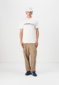 Hvit bomull t-skjorte med rosa grafisk tekst, beige cargo-bukser og marineblå sko. En lysebat cap bæres på hodet.
