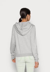 Šedá mikina vyrobená z měkké tkaniny, která má velkou kapuci, žebrované manžety a spodní lem, zobrazená zezadu s odpovídajícími světle barevnými kalhotami.