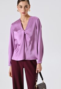 Donna che indossa una blusa di satin lavanda e pantaloni bordeaux, mentre tiene una borsa marrone scuro con rifiniture dorate.
