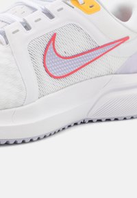 Chaussure de sport blanche avec tissu texturé gris, semelle blanche, œillet jaune et un logo Nike Swoosh violet clair bordé de rouge sur le côté.