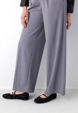 Frau trägt graue weite Hose und schwarze flache Schuhe mit Riemen, steht vor weißem Hintergrund.