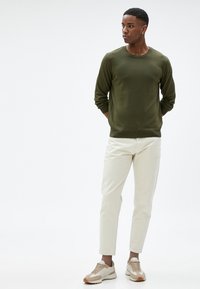 Oliwkowy sweter z dzianiny, kremowe spodnie slim-fit i beżowe sneakersy z akcentami. Luźny krój i strukturalna tkanina, postawa stojąca.