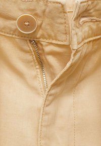 Béžové kalhoty mají skrytý zip a velký kulatý knoflík, jsou vyrobeny z měkké, texturované látky a mají viditelné švy podél švů.