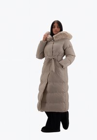 Abrigo largo acolchado beige con capucha forrada de piel, cintura ceñida con cinturón y dos bolsillos laterales. Presenta textura acolchada para aislamiento.
