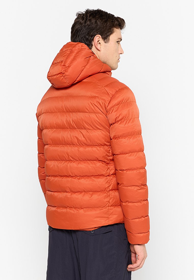 Hombre con chaqueta acolchada con capucha de color naranja brillante, de perfil, con pantalones oscuros, contra un fondo blanco liso.