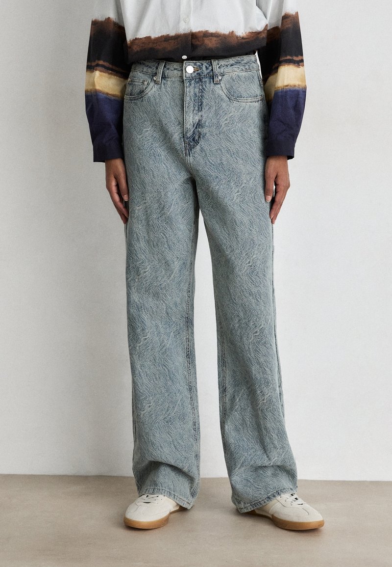 Jean bleu clair à jambes larges avec un motif texturé, avec une taille boutonnée et des poches avant, associé à des baskets blanches.