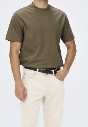 Homme portant un t-shirt à manches courtes vert olive rentré dans un jean écru avec une ceinture noire, debout avec une main dans la poche.