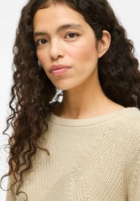 Femme aux longs cheveux bouclés et foncés, portant un pull en tricot beige, regardant avec assurance légèrement au-delà de l'objectif sur un fond blanc.