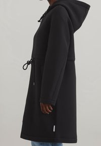 Czarny płaszcz z kapturem wykonany z gładkiego materiału. Posiada regulowany pas i kieszenie. Luźny krój i nieco wydłużony fason.
