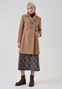 Ein brauner Wollmischmantel mit doppelreihigem Design, großen Knöpfen und einem strukturierten Kragen, kombiniert mit einem karierten Rock und braunen Stiefeletten.