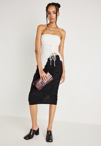 Femeie cu părul împletit, purtând un top alb fără bretele, cu volane, o fustă midi texturată neagră, mule cu tocuri închise la culoare, ținând o geantă de mână maro închis.