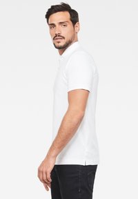 Vit pikétröja med krage och två knappar, korta ärmar, sidoflikar och mjuk tygstruktur, matchad med mörka jeans.