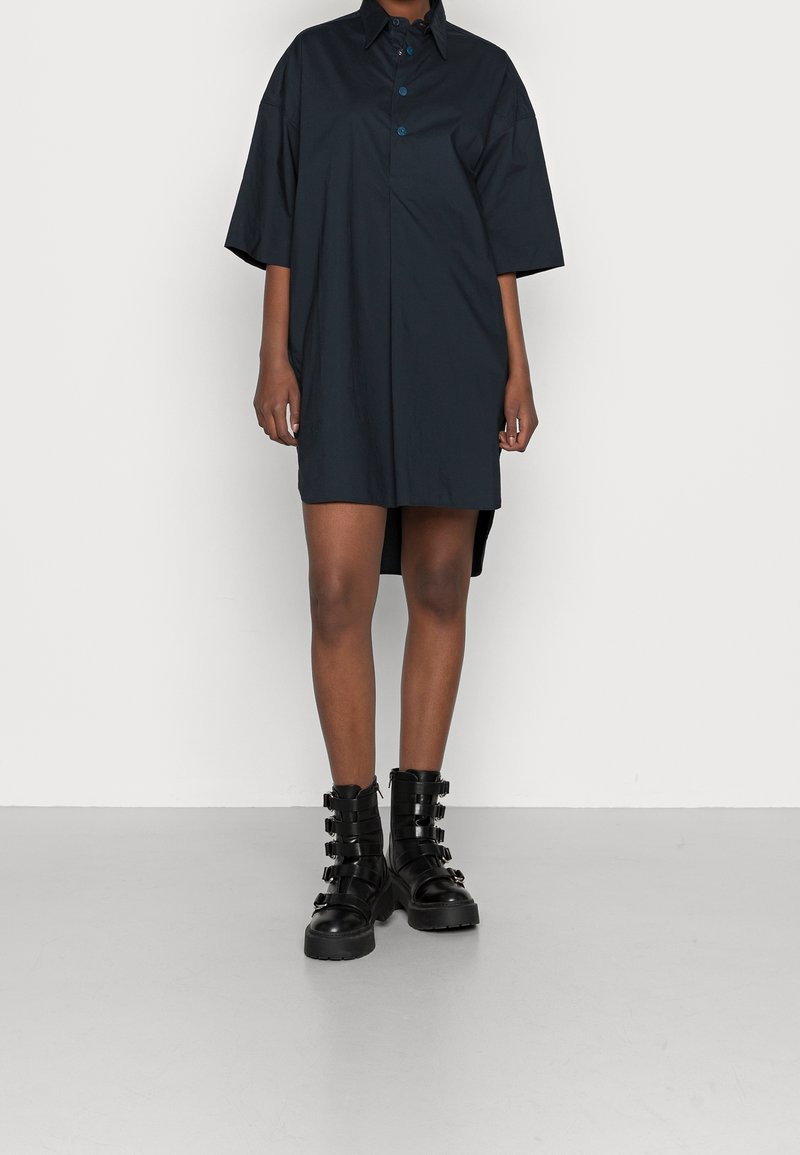 Personne portant une robe sombre ample arrivant au genou avec des manches courtes et des bottes noires épaisses à multiples boucles.
