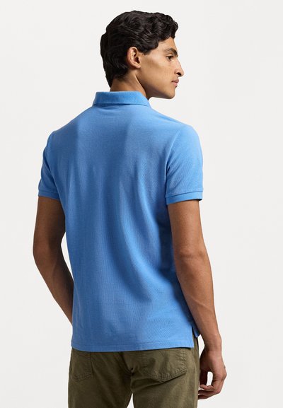 Camisa polo azul claro de manga corta, con tejido texturizado y cuello plano. Aberturas laterales destacadas en el dobladillo, combinada con pantalones verde oliva.