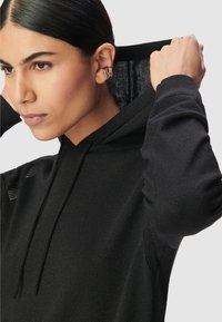 Una mujer con cabello oscuro se coloca la capucha de su suéter de punto negro sobre la cabeza, luciendo pendientes de oro en su oreja izquierda, contra un fondo liso.