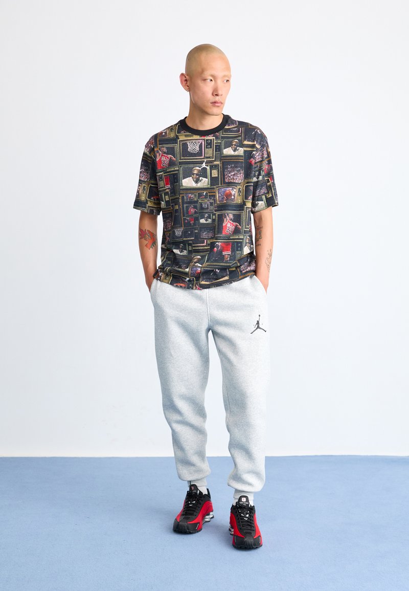 Un t-shirt graphique présente une impression multicolore de scènes de basketball. Un pantalon de survêtement gris avec un logo noir. Des baskets noires et rouges complètent la tenue.