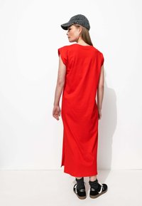 Robe rouge, mi-longue, sans manches en coton ; présente une coupe droite et des fentes sur les côtés. Associée à une casquette grise à imprimé léopard et des baskets noires.