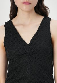 Top preto sem mangas feito de tecido texturizado com um decote em V e detalhe em nó na frente, apresentando um padrão tecido por todo o lado.