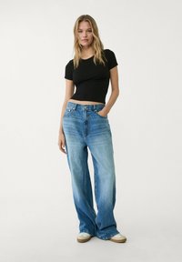 Zwarte crop top gecombineerd metlichtblauwe, wijde jeans. De jeans hebben een vervaagde wassing, vijf zakken en een relaxte pasvorm. Eenvoudige witte sneakers maken de look compleet.