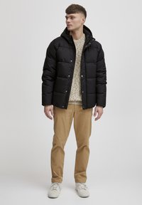 Svart boblejakke med hette og trykknapper, brukt over en beige strikkegenser, kombinert med khaki-bukser og hvite sneakers.