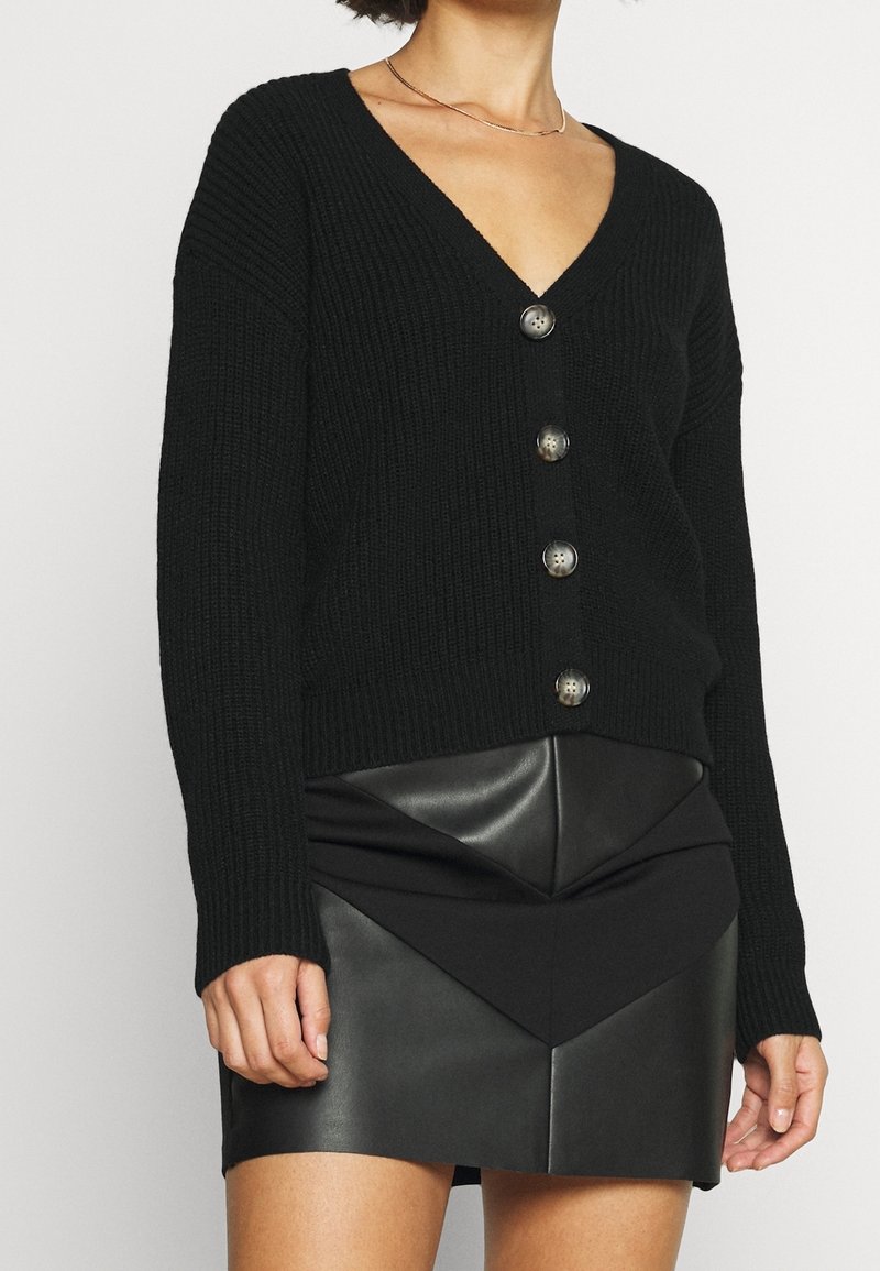 Cardigan nero a maglia con scollatura a V profonda, caratterizzato da sei bottoni rotondi lungo la parte anteriore, indossato sopra una gonna nera in finta pelle con un design geometrico.