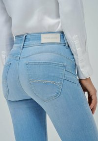 Jeans de mezclilla azul claro con una cintura de altura media, que presentan una etiqueta de 'Salsa Jeans', bolsillos traseros curvados y un detalle de costura en contraste.