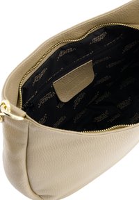 Beige leren handtas met textuur aan de binnenkant, voorzien van een zwarte voering, een gouden ritssluiting en een klein extern vak, met daarop een logo dat op stof is gedrukt.