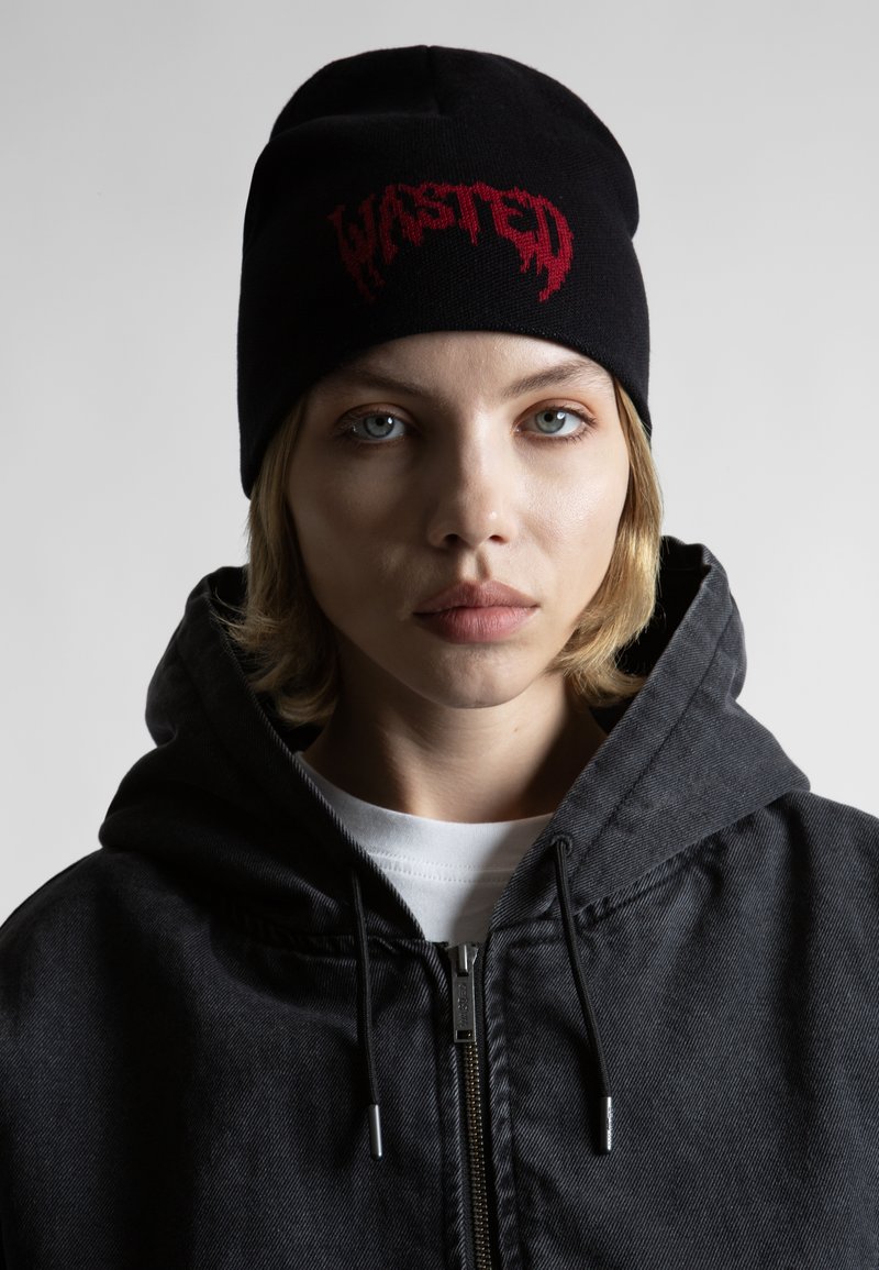 Beanie neagră cu logo-ul roșu "WASTED", purtată cu o jachetă cu glugă de culoare cărbune, dintr-un material moale. Detalii notabile includ un fermoar și șnururi.