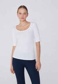 Body bianco a scollo rotondo realizzato in tessuto morbido, con maniche a tre quarti e design aderente, indossato con leggings blu navy.
