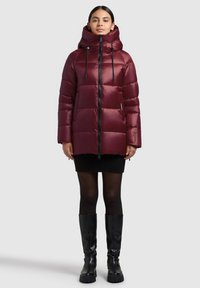 Burgundy puffer jacket with a hood, featuring horizontal quilted sections, black zipper, and drawstrings. Accompanied by a black skirt and knee-high boots.