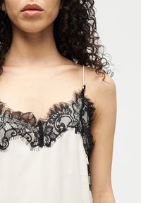 Γυναίκα που φοράει μπεζ καφτάνι με μαύρη δαντέλα στο τελείωμα και λεπτούς ιμάντες ώμου, με σγουρά σκούρα μαλλιά, ορατή από τους ώμους μέχρι τα χείλη.